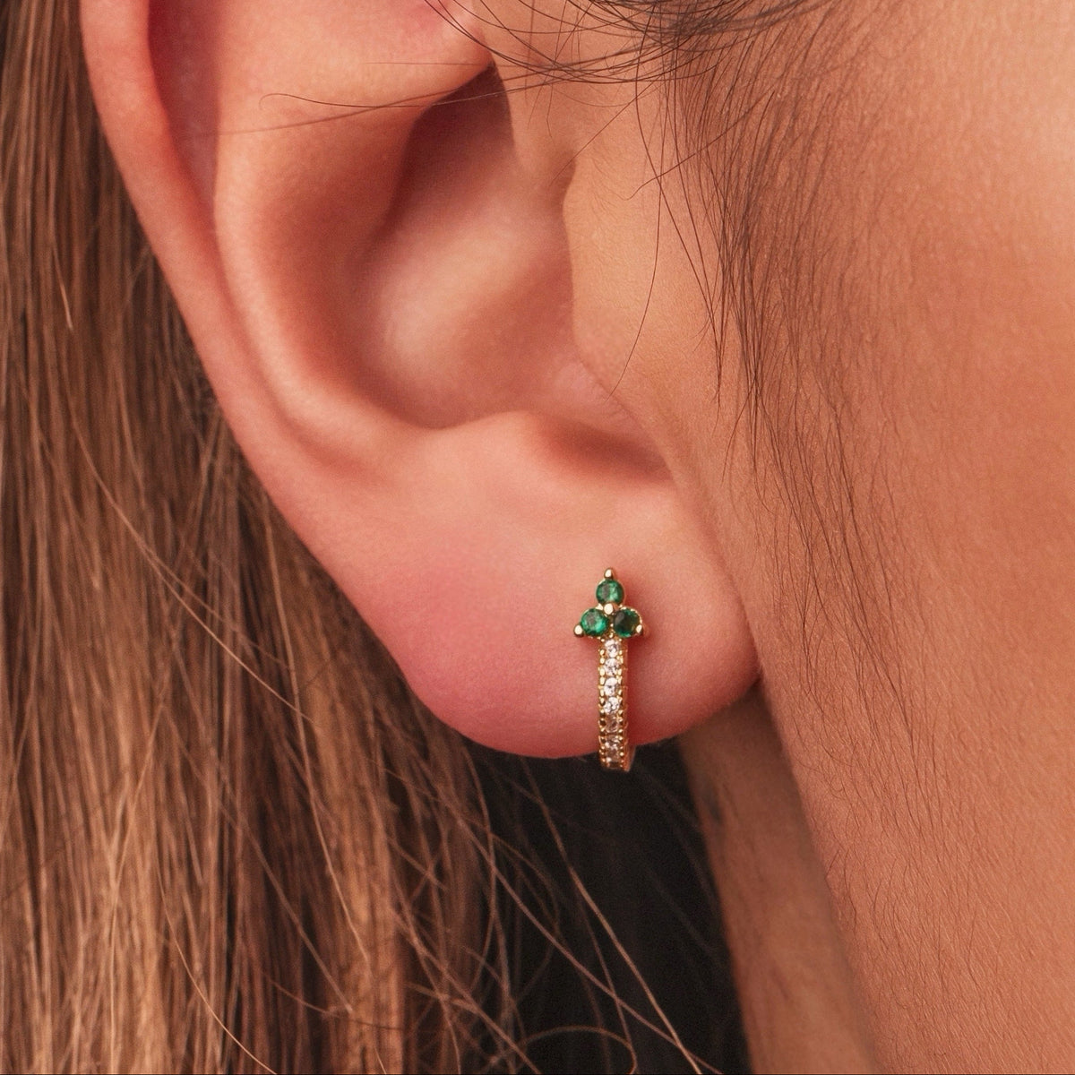 Gold-plated sterling silver huggie earrings with green and clear cubic zirconia stones worn on ear